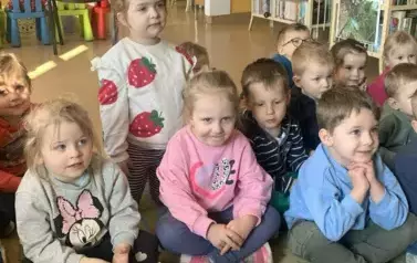 Dzieci siedzą na dywanie i z zaciekawieniem patrzą na bibliotekarkę, kt&oacute;ra czyta im bajkę.