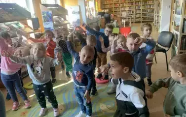Dzieci stoją w rozsypce i podnoszą ręce do g&oacute;ry naśladując Panią biblitekarkę w wykonaniu ćwiczenia.