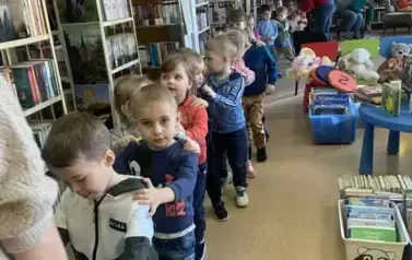 Dzieci stoją jeden za drugim gęsiego wychodząc z biblioteki. Jedna z Pań prowadzi ich z przodu, a druga stoi z tyłu na końcu kolejki.