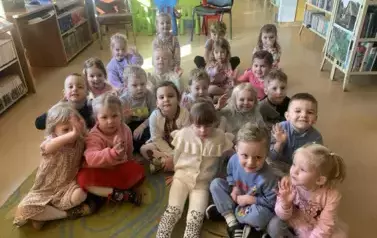 Dzieci siedzą na dywanie i słuchają bajki jaką czyta im bibliotekarka. Niekt&oacute;re z nich podnoszą rączkę w celu chęci zabrania głosu.