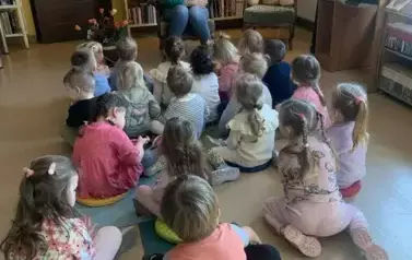 Dzieci siedzą na dywanie i siedziskach i patrzą na bibliotekarkę, kt&oacute;ra im opowiada o książce i o baśniach Andresena.