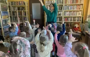 Na środku stoi bibliotekarka trzymając ręce w g&oacute;rze, dzieci stojące przed Nią robią to samo i r&oacute;wnież wyciągają ręce w g&oacute;rę.