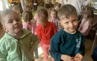 Z przodu stoi dw&oacute;ch chłopc&oacute;w, jeden z nich trzyma cukierka. W tle są inne dzieci i ich Panie.