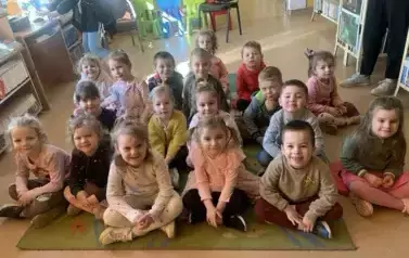 Dzieci siedzą na dywanie i słuchają bajki, kt&oacute;rą czyta im bibliotekarka. Uśmiechają się.