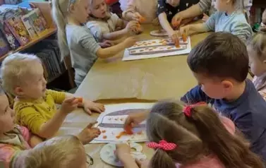 Dzieci podzielone na dwie grupy przy stole ozdabiają stempelkami z marchewki narysowany na dużych kartkach tort.