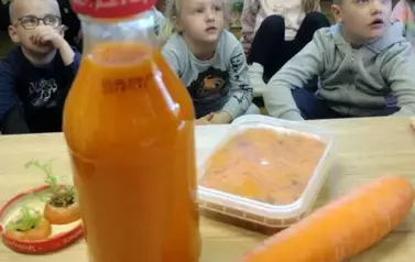 Dzieci siedzą na siedziskach, a przed nimi na biurku leżą: sok Kubuś, marchewka potarta w pojemniku, marchewka, i na nakrętce od słoika leżą dwie odcięte nacie marchewki.