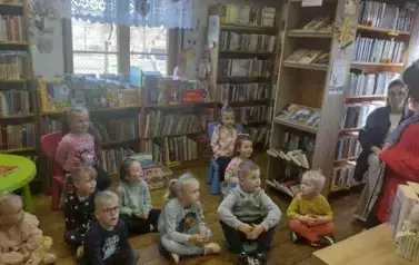 Dzieci siedzą na siedziskach i patrzą na Panią prowadzącą, kt&oacute;ra przodem do nich coś m&oacute;wi.