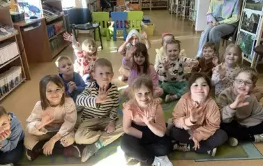 Dzieci siedzą na dywanie i siedziskach i kiwają do zdjęcia rączkami uśmiechając się.