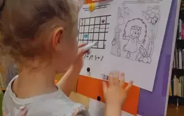 Dziewczynka w blond włosach i w kitku stoi przy tablicy na kt&oacute;rej są przywieszone zadania. Dziewczynka szuka na obrazku z Calineczką schowanych przedmiot&oacute;w kuchennych i zaznacza je pisakiem.