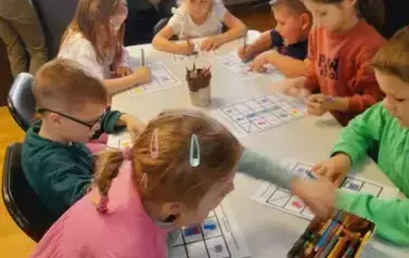 Dzieci przy białym stole uzupełniają karty pracy. Przy stole stoi ich Pani i patrzy jak to robią.