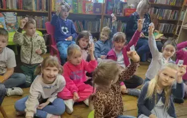 Dzieci siedzą  na siedziskach w bibliotece. Niekt&oacute;re z nich podnoszą rękę w celu zabrania głosu