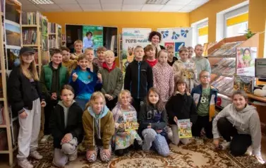 Dzieci stoją w grupie z Anną Sakowicz. Przednie dzieci siedzą na dywanie i niekt&oacute;re z nich trzymają zakpione książeczki z serii - Poprańcy