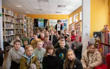 Duża grupa dzieci siedzi na dywanie,a za nimi stoi uśmiechnięta Anna Sakowicz.