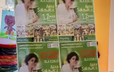 Na sztaludze wisi tablica z czterema plakatami o spotkaniu autorskim dla dzieci.