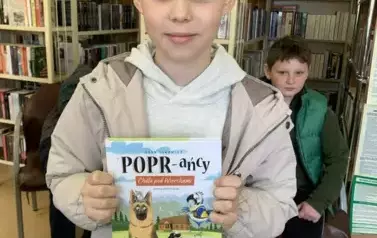 Chłopiec w blond włosach pokazuje swoją kupioną książeczkę. Za nim w tle widać z tyłu siedzącego chłopca na krześle.