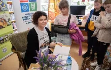 Anna Sakowicz przy stoliku podpisuje dedykację na książce, obok stoją dzieci z zamiarem kupienia książki.