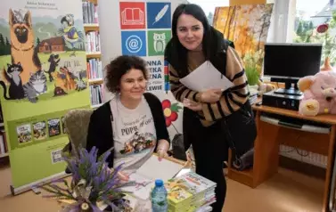 Pani w ciemnych włosach pochyla się blisko Anny Sakowicz, kt&oacute;ra siedzi przy stoliku trzymając w rękach książki. Obie Panie się uśmiechają.
