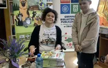 Przy okrągłym stoliku na kt&oacute;rym są książki , kwiaty i woda siedzi Anna Sakowicz. Przed Nią stoi chłopiec w blond włosach.