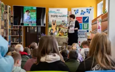 Dzieci patrzą na Annę Sakowicz, kt&oacute;ra stoi z przodu z podniesionymi lekko rękami. Za Nią duży ekran  z wyświetlanym plakatem spotkania i dwa rolapy : Poprańcy i Biblioteki w Zblewie.