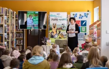 Anna Sakowicz stoi przed dziećmi i opowiada. Za Nią po lewej strone jest duży ekran z wyświetlanym plakatem spotkania, po środku rolap z bajki Poprańcy, a po prawej stronie rolap z biblioteki w Zblewie.