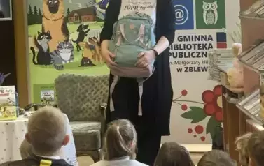 Anna Sakowicz stoi przed dziećmi trzymając plecak szkolny. Za Nią są rozwinięte dwa rolapy z Poprańc&oacute;w i Biblioteki w Zblewie.