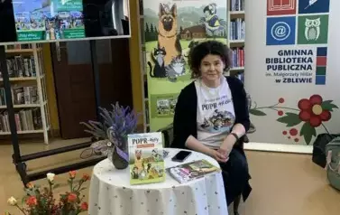 Anna Sakowicz siedzi przy okrągłym stoliku na kt&oacute;rym są fioletowe kwiaty i książki serii Poprańcy, kt&oacute;re też są ułożone na dywanie w poziomym rzędzie przed stolikiem.Z lewej strony jest duży ekran z wyświetlonym plakatem dot. spotkania.