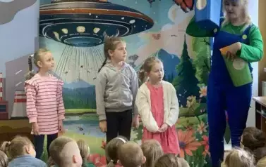Przed dziećmi stoją na środku trzy dziewczynki i aktorka trzymająca kolorowe kosze do segregacji śmieci.
