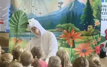 Przed publicznością dziecięcą siedzi aktor w przebraniu białego misia. W tle jest statek kosmiczny lądujący na ziemi i fruwające motyle.