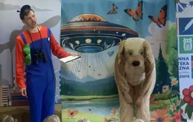 Przed dziećmi stoi pochylony aktor w przebraniu za psa, a obok aktor z lornetką i notesem w niebieskim kombinezonie. Wiszą u spodni mu zielone okulary.