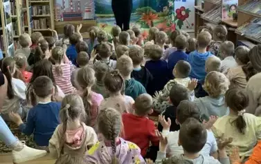 Liczna grupa dzieci patrzy do przodu gdzie stoi kobieta w białej bluzie i ma podniesione ręce do g&oacute;ry. W tle statek kosmiczny na ziemi i motyle, a z lewej strony zadymione miasto.