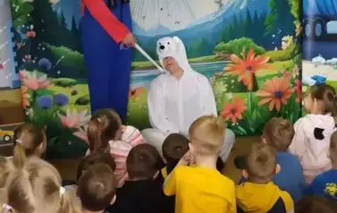 Przed dziećmi siedzi aktor przebrany za białego misia, a obok stoi drugi aktor w niebieskim kombinezonie i trzyma misia za ogon. W tle statek kosmiczny lądujący na ziemię.