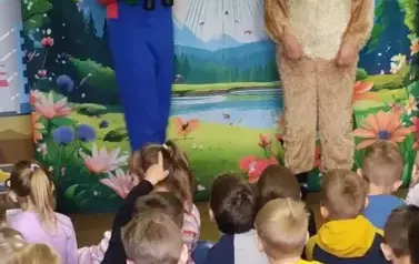 Przed dziećmi stoi przebrany piesek, a obok aktor w niebieskim kombinezonie, kt&oacute;ry trzyma notes i zielone okulary, na szyi wisi mu lornetka. W tle statek kosmiczny lądujący na ziemi.