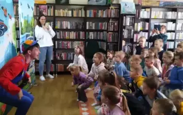 Dzieci z publiczności stoją  w p&oacute;łprzysiadzie naśladując aktora, kt&oacute;ry im to pokazuje.