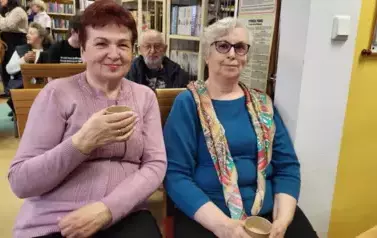 Dwie starsze Panie siedzą na krzesłach i piją kawkę. Uśmiechają się.