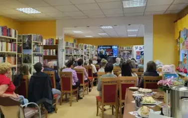 Widok od tyłu na całą bibliotekę wypełnioną Czytelnikami, z przodu autor opowiada o książce