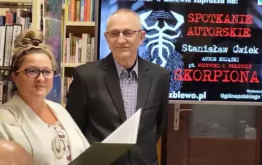 Emilia Bielska wita Stanisława Ćwieka, kt&oacute;ry stoi obok. Czyta informacje o nim. Za nimi ekran duży z wyświetlanym plakatem dotyczącym tego spotkania. 