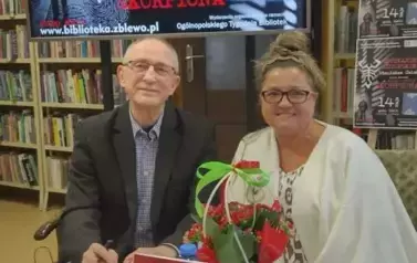 Przy okrągłym stoliku siedzi kierowniczka biblioteki z Panem Stanisławem. Uśmiechają się. Na stoliku jest kwiatek w koszyczku, woda i  książka Pana Ćwieka.