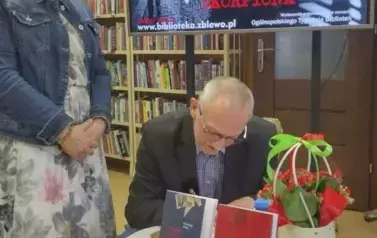 Czytelniczka w blond włosach i okularach stoi przy stoliku gdzie Pan Ćwiek podpisuje dedykację dla Niej na nowo zakupionej książce.