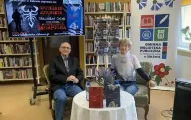 Przy okrągłym stoliku z kwiatkami i wystawionymi książkami siedzą autor książki i Jego prowadząca. Z lewej strony wyświetla się na ekranie plakat o tym spotkaniu, po środku jest sztaluga z czterema plakatami ,a po prawej stronie rolap biblioteki w Zblewie