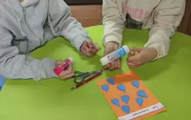 Dwie dziewczynki siedzą przy okrągłym zielonym stoliku gdzie leżą kleje, nożyczki i pomarańczowa kartka z niebieskimi elementami w kształcie stożka z naklejonym tekstem.