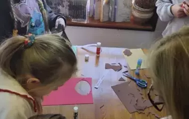 Przy brązowym stole stoją dzieci i wycinają coś z kolorowego papieru i przyklejają na kartki.