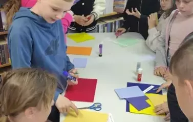 Przy białym dużym stole stoją dzieci i każdy z nich ma kolorową kartkę przy sobie, klej, nożyczki i pasek z wydrukowanym tekstem.