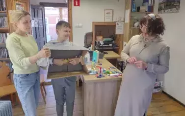 Chłopiec trzyma na piersiach karton w kt&oacute;rym ma włożone ręce i coś w nich trzyma. Obok stoi nauczycielka i Pani prowadząca zajecia. Chłopiec musi odgadnąc co trzyma w dłoniach.