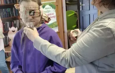 Dziewczynka w blond włosach i w fioletowym dresie stoi z zawiązanymi szalem oczami. Pani prowadząca zajęcia podaje jej pod nos słoiczek do wąchania.