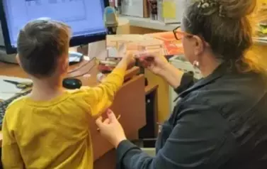 Bibliotekarka siedzi przy komputerze, a przy Niej stoi chłopiec z książką w ręku i bibliotekarka pomaga mu przyłożyć tą książke do czytnika.
