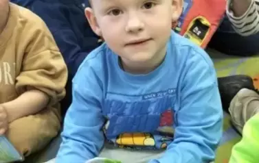 Chłopiec w blond włosach i w niebieskiej bluzce bawi się książeczką pluszową, obok są inne dzieci  bawiące się książeczkami i ich Pani.