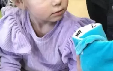 Dziewczynka w blond włosach i w fioletowej bluzce trzyma w rączce niebieską pluszową książeczkę. W tle widać inne dzieci.