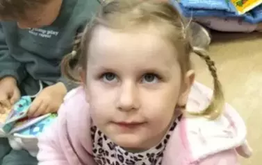 Dziewczynka w blond włosach i malutkich warkoczykach przegląda pluszową książeczkę o biedronce. Obok widać inne dzieci z książeczkami.