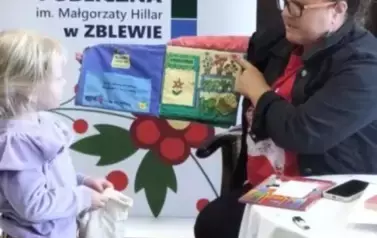 Przy stoliku siedzi bibliotekarka z otwartą pluszową książeczką o sklepie. Przy niej stoi dziewczynka w blond włosach z malutką siateczką  bawiąc się w robienie zakup&oacute;w odklejając z książeczki r&oacute;żne elementy przedtawiające żywność.