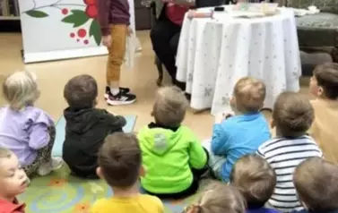Dzieci siedzą na siedziskach w bibliotece, z przodu przy stoliku siedzi bibliotekarka z książeczką pluszową w rękach, a przy Niej stoi chłopiec trzymając malutką siateczkę do zabawy w robienie zakup&oacute;w.